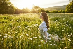a beautiful woman in a light dress sits in a chamomile field against the backdrop of the setting sun and enjoys nature