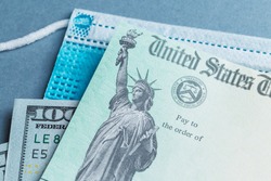 A US stimulus check on with some one-hundred-dollar bills and a medical mask