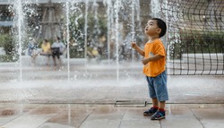 An adorable toddler Asian boy (1-year-old) standing and enjoy to see outdoor public fountains a water splashing for the first time. Thailand plays and fun space for kids. Vacation and holiday concept.