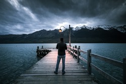Unknown man  standing on wooden bridge by the shore of Wakatipu lake at Glenorchy in south island of Newzealand during cloudy night with dim light from light bulb on the bridge