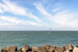 The lighthouse with heliplatform at the entrance of the Port of Rotterdam in the Netherlands. There are concrete wave breakers in the foreground and in the blue sky above are white cirrus clouds.