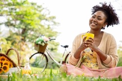 Smiling charming African American woman in imagined dreaming with favorite beverage after hard working last holiday during the picnic on the sunny meadow. Relax and Leisure concept.