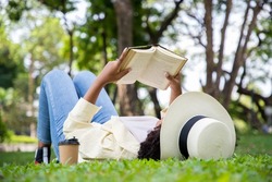 Side view of charming woman reading book in grass under tree with cup of coffee. Relax in summer time holiday laying on the grass field comfortable feeling.
