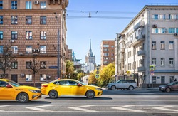 View of the Ukraine hotel between two houses on Smolenskaya Square in Moscow and a yellow taxi on the highway on a summer sunny day. Caption: Smolenskaya Square 13/21