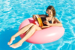 Young happy girl in bikini is swimming in the pool and reading a book with a pink circle.