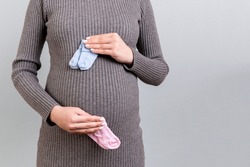 Close up of pregnant woman in gray dress holding baby pink and blue socks against her belly at gray background. Expecting twins. Is it a boy or a girl? Child expecting concept. Copy space.