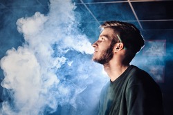 The guy smokes, exhales smoke. Steam from an electronic cigarette. Portrait of a young man in the smoke. Blows steam vortexes by hand with spots of light on the face