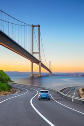 Cars in motion in the highway landscape under the Bosphorus bridge. Road landscape at colorful sunset. Car driving on the road. Nature scenery on city beach. Travel journey for summer trip on road.