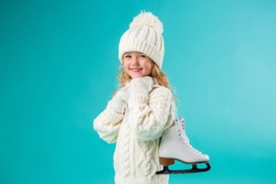 little blonde girl smiling in a winter white hat and sweater, holding skates. isolate on blue background, space for text. winter sports