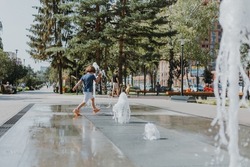 little boy is running in the street and playing with the water jets of a fountain spouting from the ground. child in blue T-shirt and white shorts is fooling around outdoors. lifestyle. space for text