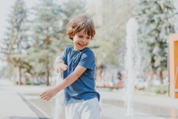 little boy is running in the street and playing with the water jets of a fountain spouting from the ground. child in blue T-shirt and white shorts is fooling around outdoors. lifestyle. space for text