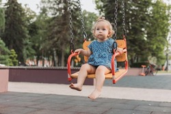 sweet baby in a blue dress swings on a swing on a playground in the park. child walks outdoors. lifestyle. space for text. High quality photo