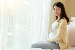 Happy pregnant young asian woman holding belly and look at camera with happiness smiley face. Young Asia pregnancy mother love her baby and has good health. Mom waiting, expecting baby has good health