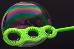 A bubble stick, with three holes of various sizes from which to blow bubbles, with an iridescent bubble straddling all three. 