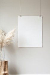 Vertical hanging poster mockup on a neutral coloured wall. Hanging by clips and string. Pampas grass in a large glass vase sitting on a small tree stump prop.