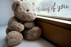 The sad teddy bear sits on the windowsill and looks sadly out the window, it's raining, the glass is covered with drops. Handwritten inscription on a glass 