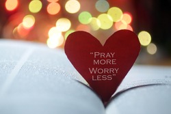 Inspirational quote on a red heart - Pray more, worry less. Spiritual text message on an open bible book page with colorful bokeh lights background. Faith, hope, love and believe in God concept.