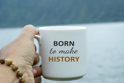 Inspirational words on a cup of coffee or tea - Born to make history. With closeup of a person holding white coffee cup on blue lake nature background. Life purpose concept.