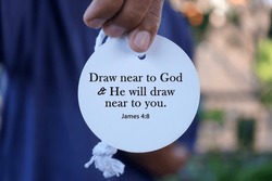 Bible verse quote - Draw near to God and He will draw near to  you. James 4:8 with person showing text on white circle label paper in hand. Christianity concept with bible verses quote.