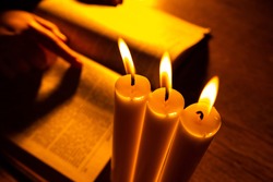 Candle church close up. candle church next to bible. Book as a Symbol of Religious Literature. Blurred hands on bible in the background. A man reads bible next to a candle church. Praying hands