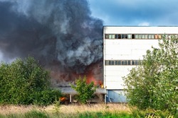 Fire next to industrial building. Black smoke from fire. Fire in factory or factory. Black smoke on blue sky background. Concept is industrial accident. Flame next to industrial building.