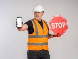 Red stop sign and smartphone in builder's hand. Man in reflective vest demonstrates smartphone with place for text on the screen. Forbidding sign. Prohibition of construction. Restrictive measures.