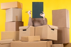 Woman entrepreneur. Girl among cardboard boxes. Boxes as business metaphor. Owner of online store concept. Woman closes face clipboard. Workaholic girl on yellow. Businesswoman has lot of work