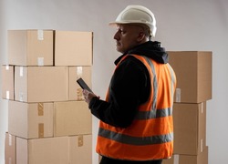 Postal warehouse worker. Experienced man with smartphone. Guy in orange vest is in mail warehouse. Postal company warehouse with boxes. Cardboard parcel on gray. Career in courier company