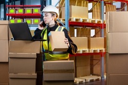 Postal warehouse. Woman works in delivery service warehouse. Storekeeper service courier company on phone. Girl works in warehouse of postal service. Woman in yellow vest looking at laptop