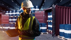 Man in fuel depot. Barrels with fuel in shipping containers. Guy logistician works in oil industry. Human in yellow vest holds laptop. Preparation of fuel for import concept. Export of oil products
