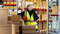 Postal warehouse. Woman in protective helmet is talking on phone. Girl in yellow vest next to boxes. Workflow in warehouse of postal service. Shelving with pallets and boxes behind woman.