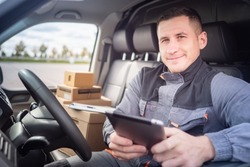 logistic delivery of something. Courier is engaged in logistic delivery. Courier driving minivan. Postman in gray uniform. Parcel cardboard boxes behind postman. Courier smiling looking at camera
