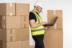 Postal worker with parcels. Postal worker in headphones with microphone. Man in yellow vest and hardhat. Postal worker is holding clipboard. Corton boxes for delivery to client. Delivery business