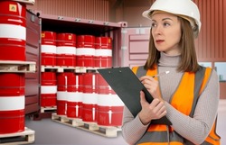 Warehouse worker woman. Warehouse of chemical company. Girl in orange vest is holding clipboard. Red barrel of chemical products. Chemistry barrels in blurred maritime container. Chemical industry