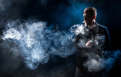 Man in smoke from vaping. Unrecognizable vaper guy. Human vaping while standing in dark. Vaper blows steam out nose. Man with electronic gadget for vaping. E-cig in hand male. Electronic cigarettes