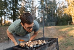 Middle age caucasian man blowing air into the bbq coals while cooking grilled champignons. Outdoors BBQ party, simple tasty foood on fire