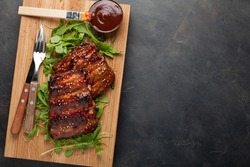 Closeup of pork ribs grilled with BBQ sauce and caramelized in honey on a bed of arugula. Tasty snack to beer on a wooden Board for filing on dark concrete background. Top view with copy space.