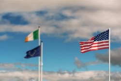 Waving flag of United States of America against pastel light blue color sky in focus. National flag of Ireland and Euro Union out of focus. International relationship theme