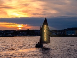 Silhouette of a boat sailing into town. Glowing sign 2023 on the sail of the yacht. Celebrating new year concept. Dramatic cloudy sky in the background with rising sun.
