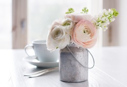 Little delicate elegant bouquet of flowers, buttercups and white lilacs in tin can and a teacup on white wooden table, morning breakfast