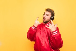 Expressive man with a beard listens to music in the headphones, wears a red jacket, stands on a yellow background. Young man listens to rock music and shows a heavy metal gesture. Isolated.