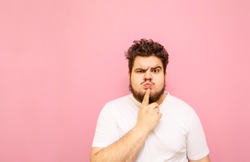 Portrait of a funny curly overweight man in a white t-shirt stands on a pink background and looks into the camera with a worried pensive face. Charismatic fat man looking dissatisfied at the camera.
