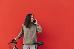 Happy woman in casual clothes and with a bicycle communicates on the phone on a red background, looking aside at smiling. Isolated. Copy space
