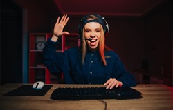 Positive female streamer sits at night in the room at the computer and communicates on a headset with subscribers, poses palms of greetings to the camera and smiles.