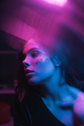 Fashionable photo of a woman with bright makeup in neon light with purple and blue in a dark room, looking away. vertical