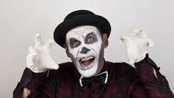 A horrible man in clown makeup grimaces and makes frightening gestures. A scary clown looks at the camera and laughs terribly.