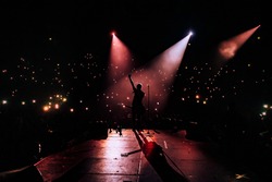 Music show. Guitarist in front of crowd on scene in night club. Bright stage lighting, crowded dance floor. Phone lights at concert. Band red silhouette crowd. People with cell phone 