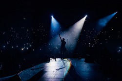View from the stage stadium. Vocalist of a popular pop band on the background of the flashlights  of phones during a concert. Fans switch on lights on smart phones on tribunes. Crowd waving cellphones