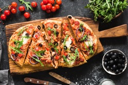 Flatbread pizza garnished with fresh arugula on wooden pizza board, top view. Dark stone background