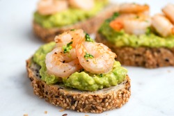 Healthy appetizer or snack avocado shrimp bruschetta with whole grain bread on white marble, closeup view. Fried shrimp and mashed avocado on a toasted bread